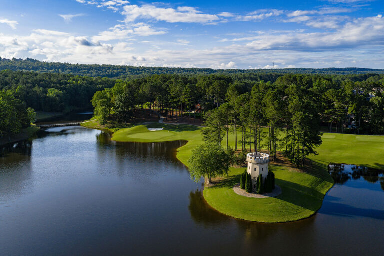 An aerial view of the Ballantrae Golf Club in Pelham, Alabama, featuring the iconic white castle tower on a green peninsula, surrounded by a calm lake and the rolling hills of Oak Mountain.