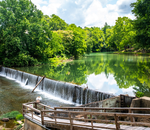A view from Old Town Helena of the Buck Creek dam and waterfall in Helena, Alabama, featuring a wooden overlook boardwalk in the foreground and lush green trees reflecting on the calm river under a cloudy sky.