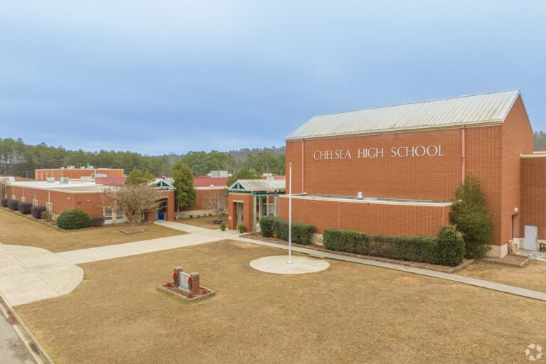 An aerial exterior view of Chelsea High School in Chelsea, Alabama, showing the brick building structures, open courtyard pathways, and landscaping under an overcast sky.