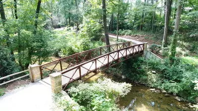A wooden footbridge crossing a shallow stream along the Jemison Park Nature Trail in Mountain Brook, Alabama, surrounded by lush green trees and forest foliage.