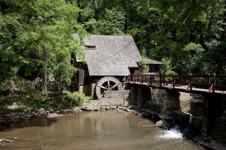 The historic Old Mill on Shades Creek in Mountain Brook, Alabama, featuring a rustic wooden water wheel and bridge surrounded by lush green forest.