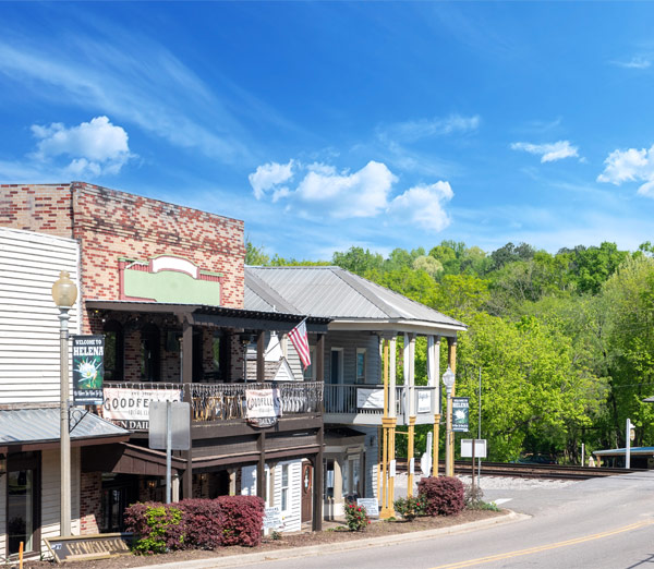 The historic storefronts of Old Town Helena, Alabama, featuring classic brick and wood architecture, two-story balconies, and a view of the adjacent railroad tracks and green trees under a bright blue sky.